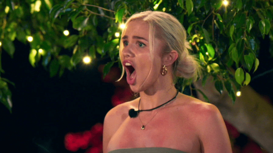 A woman with blonde hair in a bun screams in shock, her mouth wide open, wearing a strapless top and a necklace with a microphone attached.