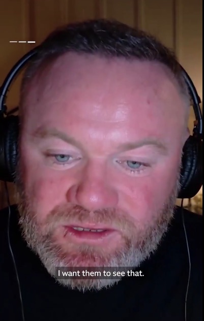 Close-up of a man with blue eyes and a beard, wearing headphones, looking down and speaking.