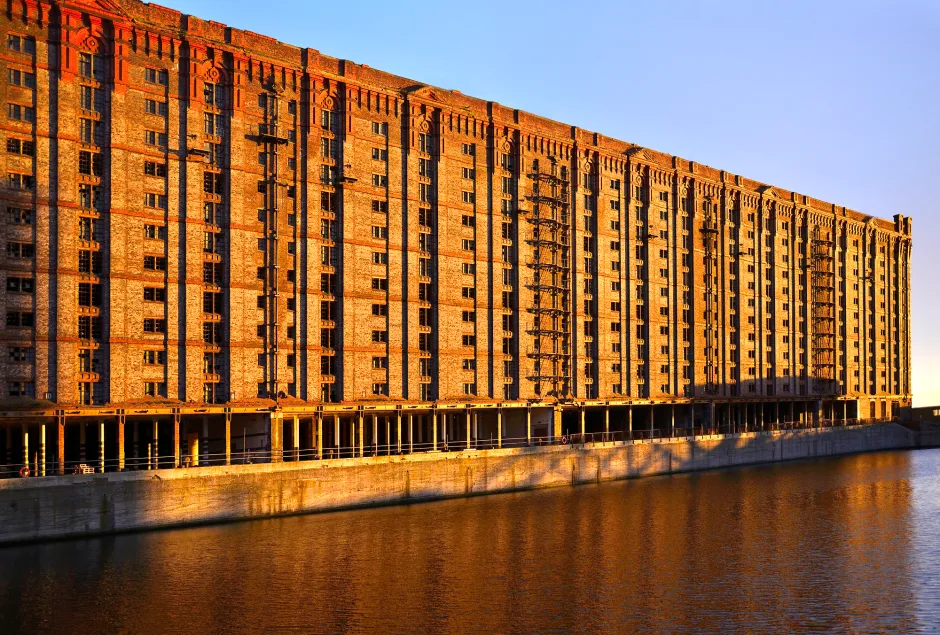 A large, long brick building, possibly a warehouse, with many small windows, stands beside a body of water under a clear sky.