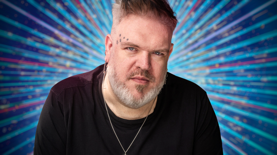 A male with a light beard, gray hair, and tattoos above his left eyebrow looks directly at the camera.