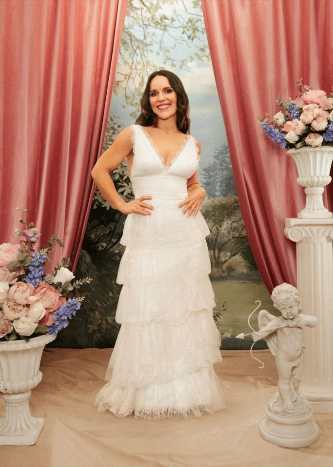 A smiling woman in a tiered white lace gown standing in a whimsical setting with pink curtains and a backdrop of a painted landscape.