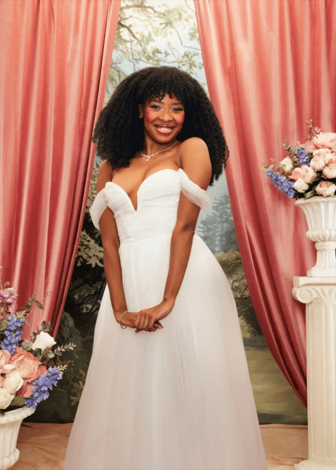 A smiling woman in a white gown with a deep V-neck, off-the-shoulder sleeves, and full skirt, standing between red curtains and floral arrangements.
