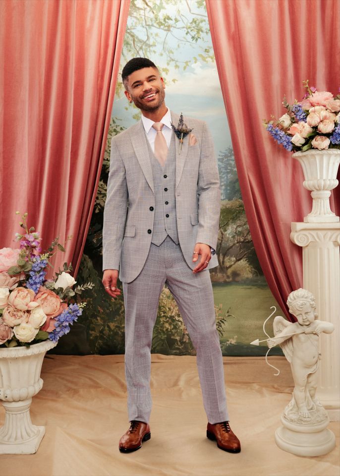 Keye in a light grey checked three-piece suit with a pink tie and a purple boutonnière.