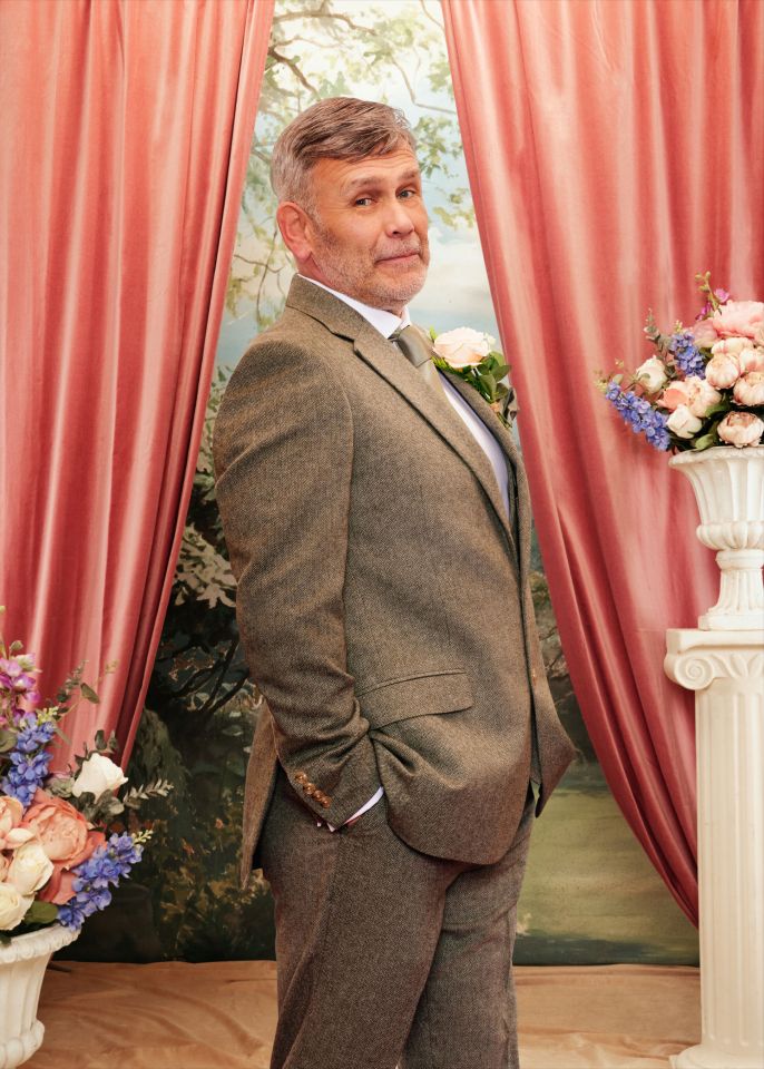 Paul in a brown tweed suit with a boutonnière, standing in front of a painted backdrop of a landscape and pink curtains.