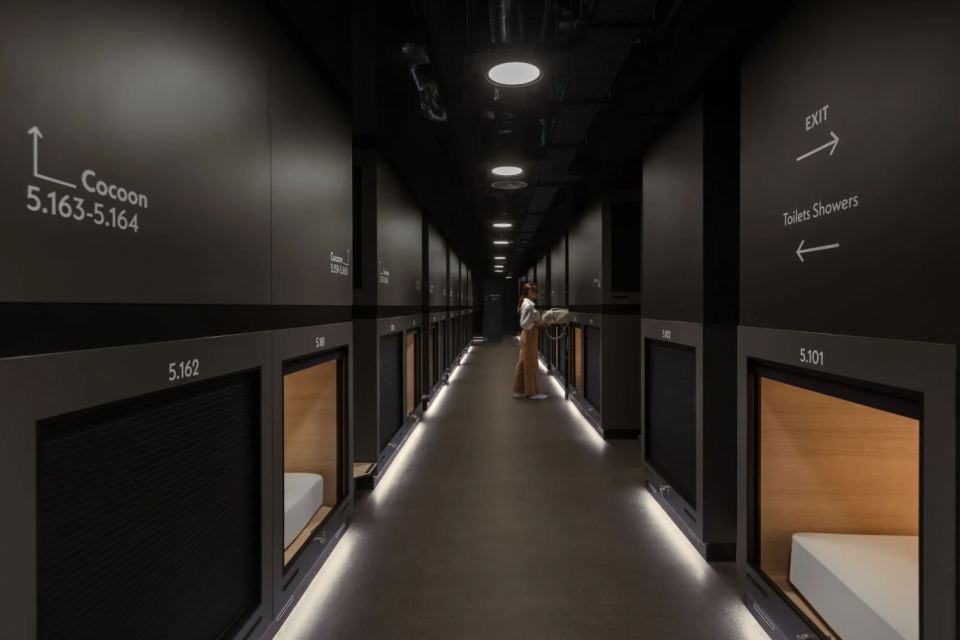 Interior view of a Zedwell Capsule Hotel hallway with individual sleep pods and a woman.
