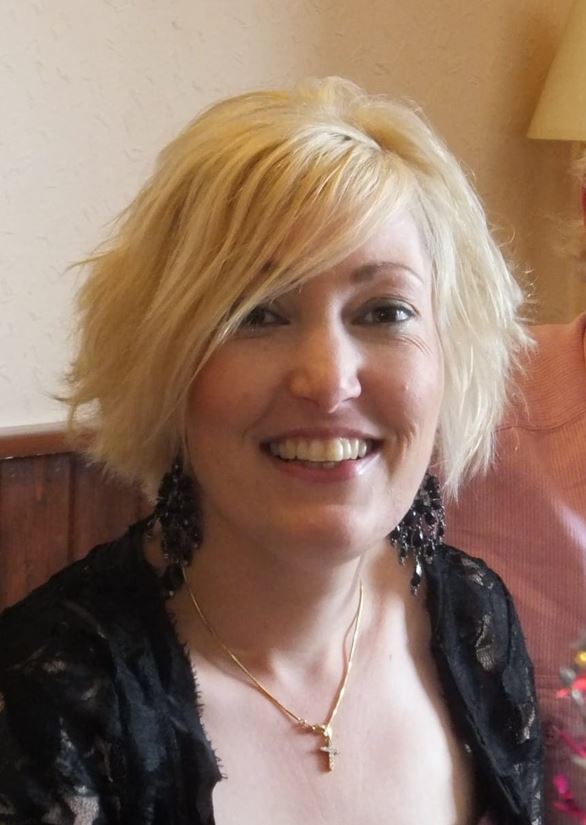 A smiling woman with short blonde hair and large dark earrings, wearing a gold cross necklace.