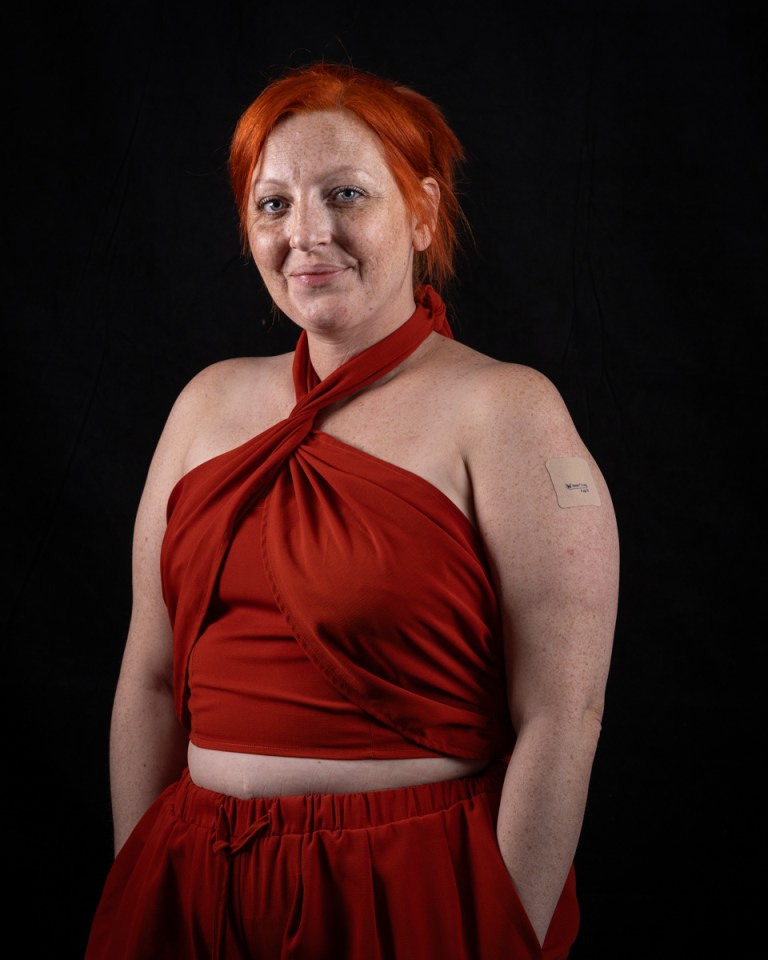 A woman with red hair and freckles smiles, wearing a red outfit, with a medical patch on her left arm.