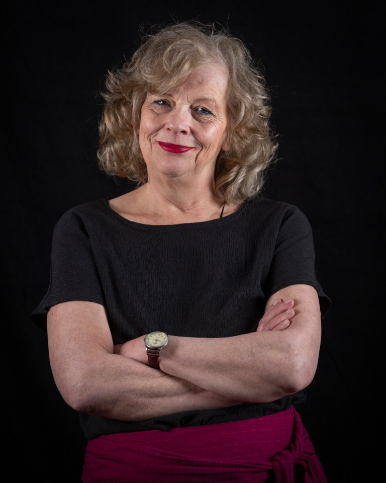 A woman with curly blonde hair and red lipstick smiling, with her arms crossed.
