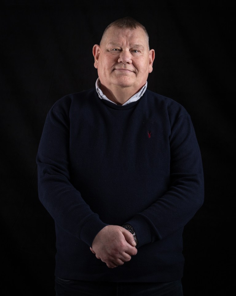 A man standing against a black background with his hands clasped in front of him.