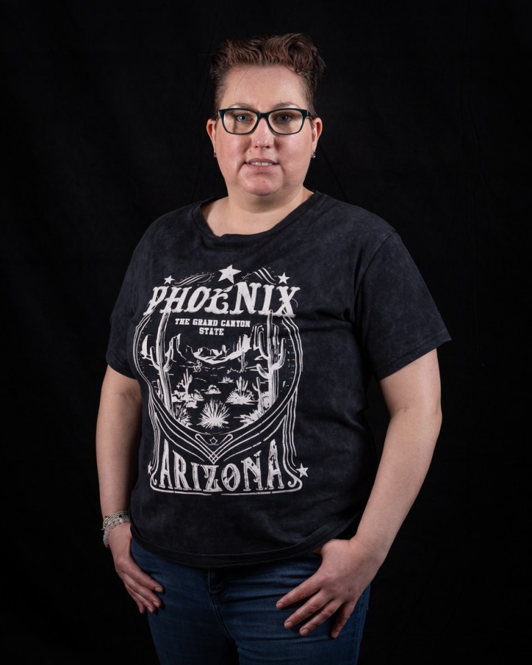 Woman standing with her hands in her pockets, wearing a black t-shirt with "Phoenix Arizona" on it.