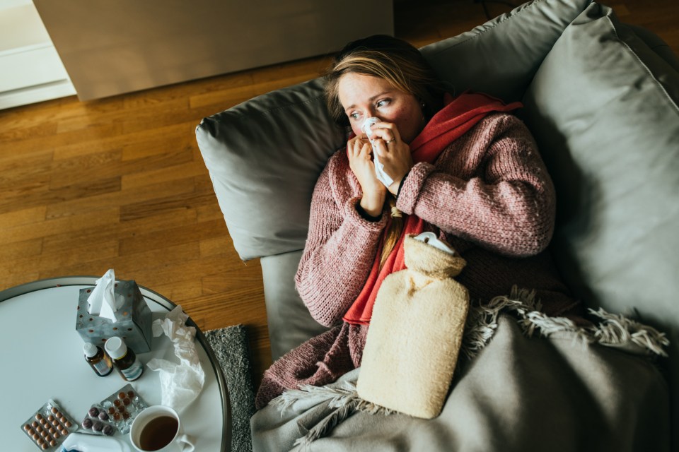 Woman lying on a sofa, blowing her nose, with a hot water bottle and medicine nearby.