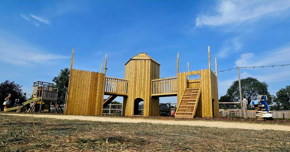 Wooden castle playground with slides and swings under a blue sky.