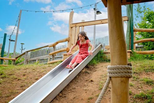 A girl in a red dress slides down a metal slide at Jack's Yard.