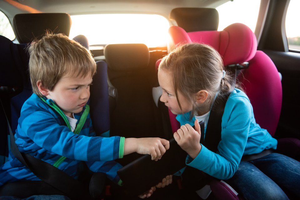 Two children in car seats in the back of a car fighting over a tablet.