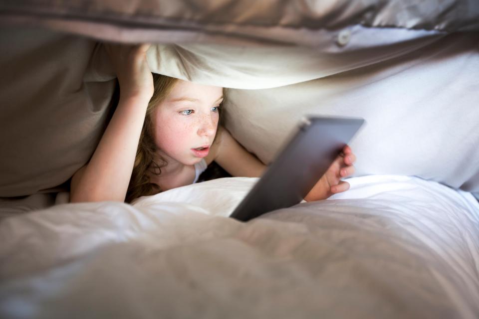 A young girl with fair skin and freckles looks at a tablet under a blanket.