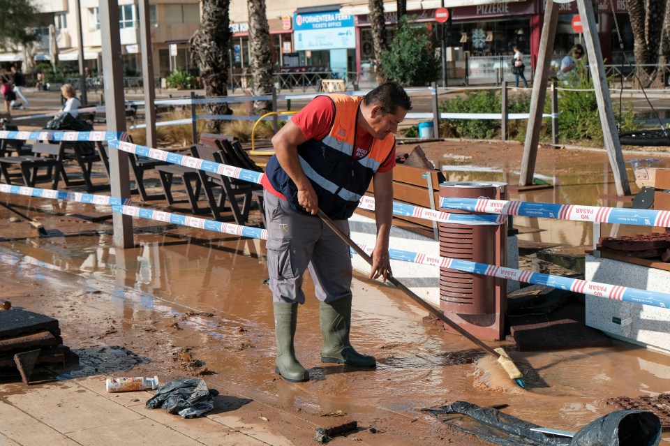 A man in a safety vest and rubber boots uses a broom to clear floodwater from a street in Ibiza, Spain.