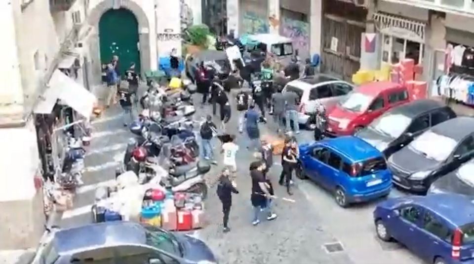 A street brawl among football fans, some throwing chairs, with cars and scooters parked on either side.