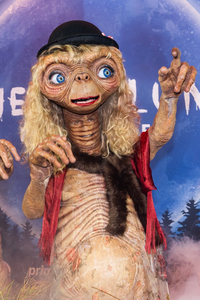 Heidi Klum dressed as an E.T. alien in a red vest and black hat, with another person's face on her chest.