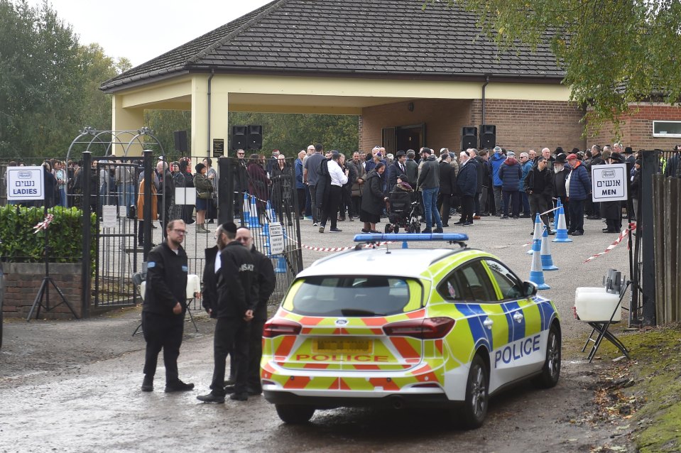 People gathered outside a synagogue for a funeral, with signs separating men and women, and a police car present.