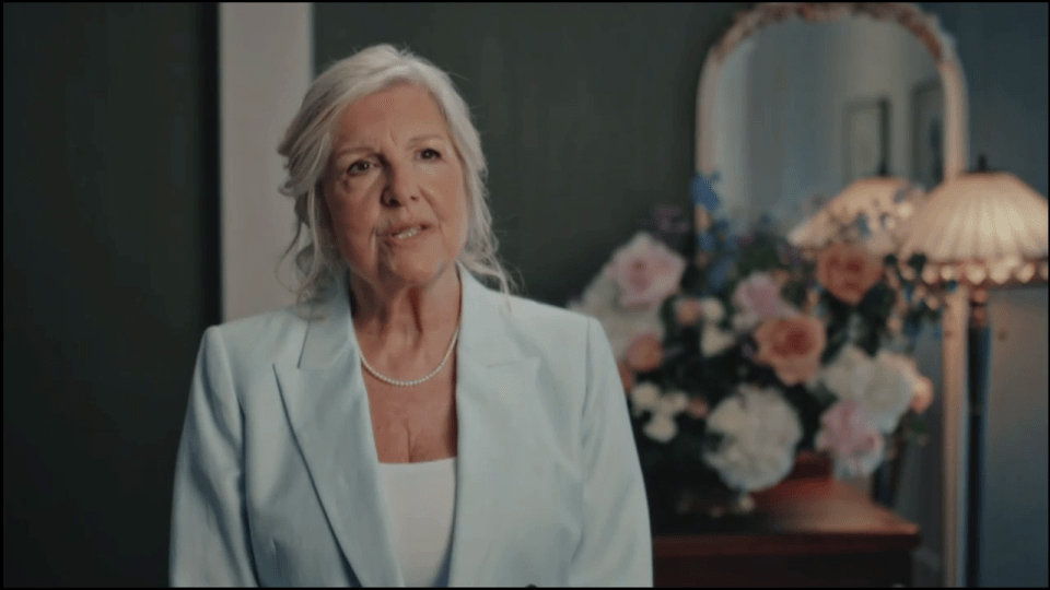 A woman with white hair and a pearl necklace speaking, with a mirror, flowers, and a lamp in the background.