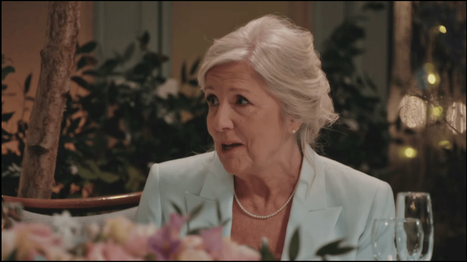 A woman with gray hair wearing a light blue blazer and a pearl necklace with a surprised expression.