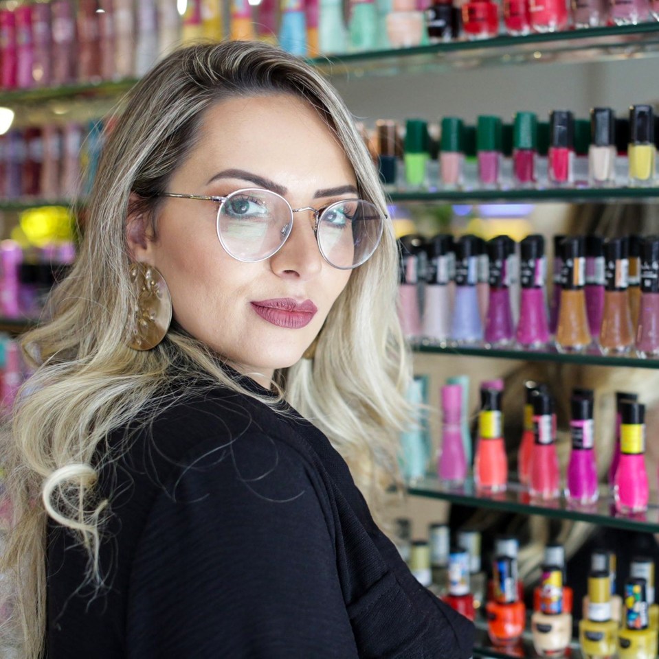 Ana Carolina Silva, a woman with long blonde hair, glasses, and dark lipstick, looking over her shoulder with shelves of colorful nail polish in the background.