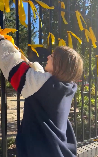 A person tying a yellow ribbon to a black metal fence.