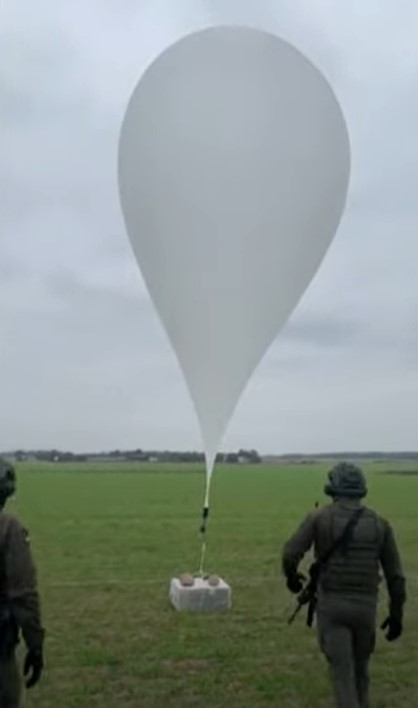 Two armed men in tactical gear standing next to a large white balloon attached to a concrete block in a grassy field.