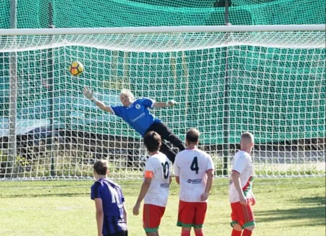 Soccer goalie Lamberto Boranga in a blue shirt diving to block a yellow and black ball.