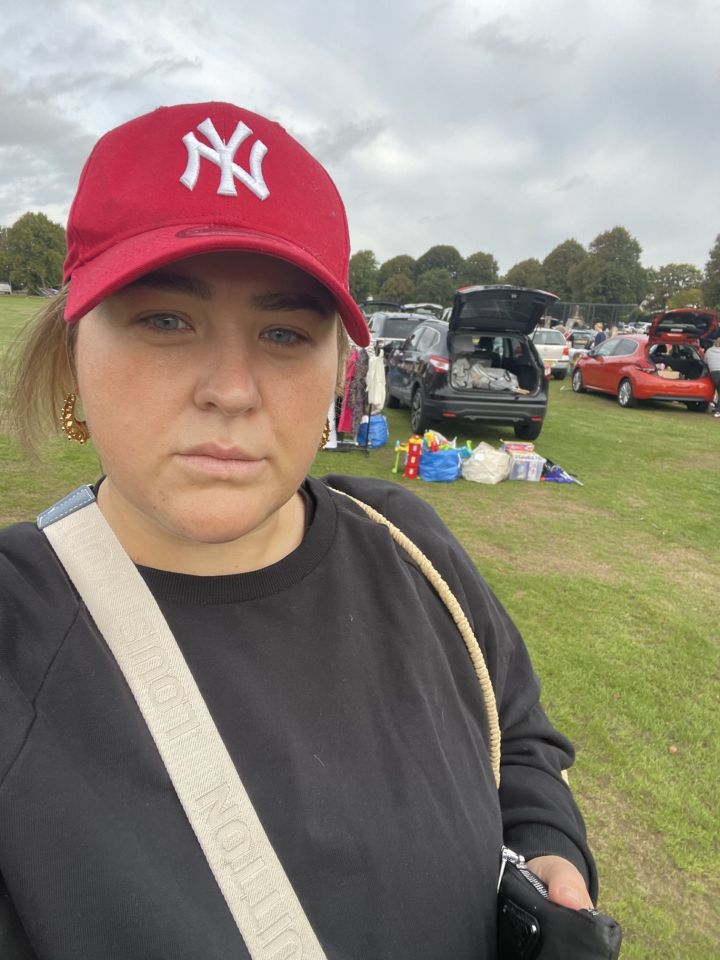 A woman in a red New York Yankees baseball cap and black sweatshirt poses for a selfie at a car boot sale.