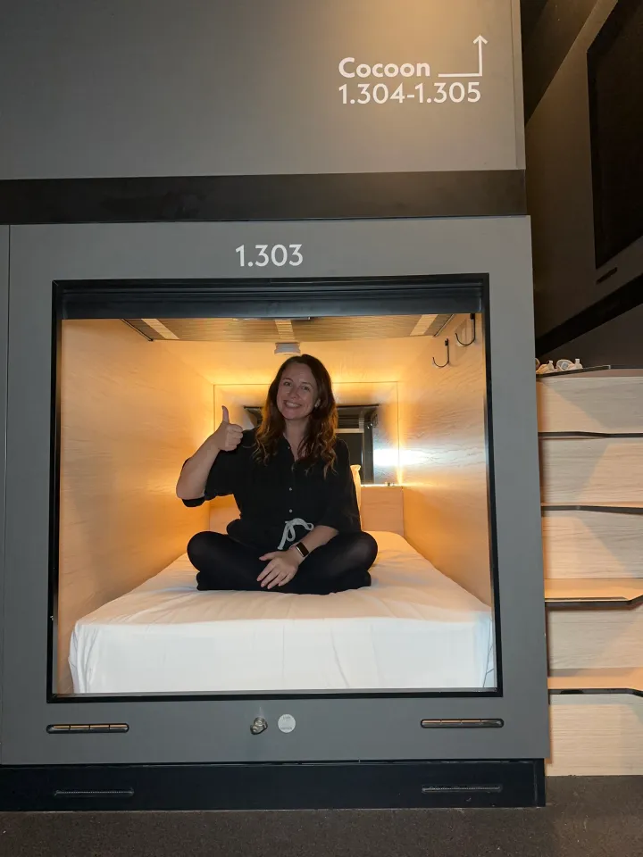 A woman gives a thumbs-up from inside a sleeping capsule, indicating satisfaction.