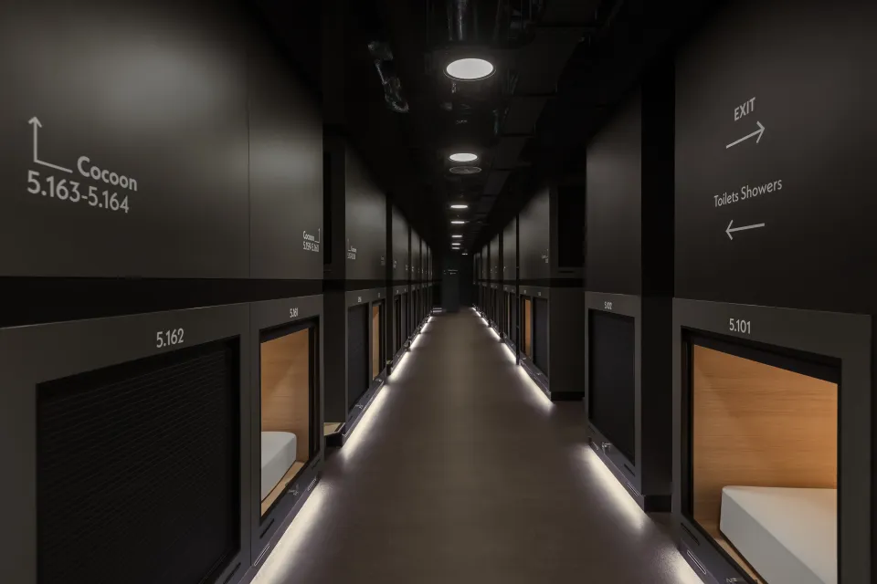 A long hallway with capsule rooms, some with open doors revealing beds, and signs for "Cocoon" rooms and "Toilets Showers" with arrows.
