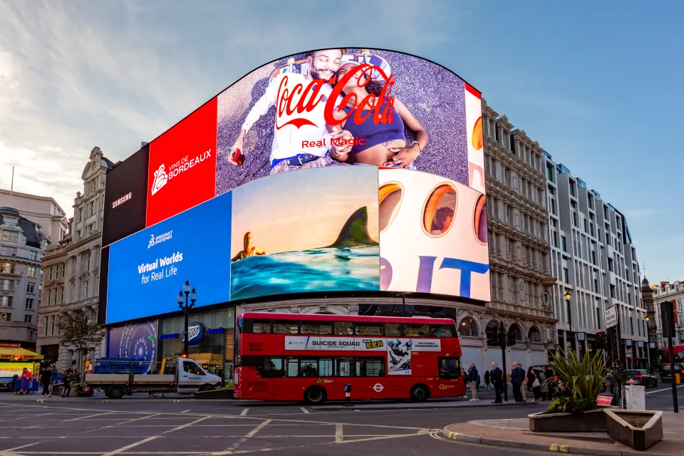 Advertising screens at Piccadilly Circus featuring ads for Coca-Cola, Vins de Bordeaux, and Dassault Systèmes, with a red double-decker bus on the street.