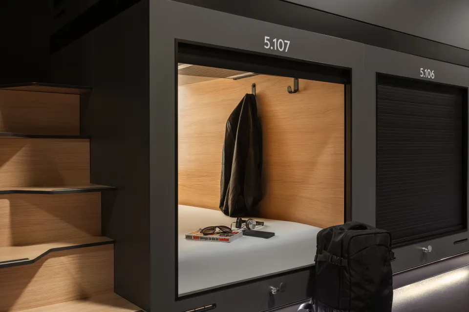An open capsule hotel room with a jacket hanging, and a book and sunglasses on the bed.
