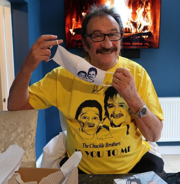 Paul Chuckle holding a face mask with his and his brother's faces on it, wearing a yellow t-shirt with the same image.