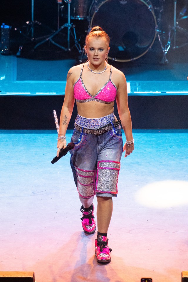 JoJo Siwa walking at America's Got Talent Studios, wearing a denim shirt, blue shorts, and white and gold boots, next to a rack of costumes.