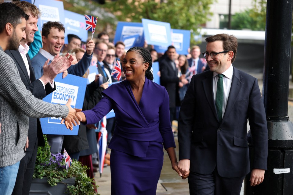 Kemi Badenoch, in a purple dress, shakes hands with a man while holding hands with her husband, Hamish Badenoch, at the Conservative Party conference.