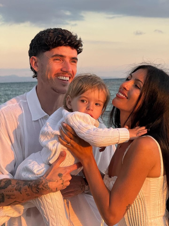 A man and woman smiling at each other while holding a baby, against a sunset ocean backdrop.