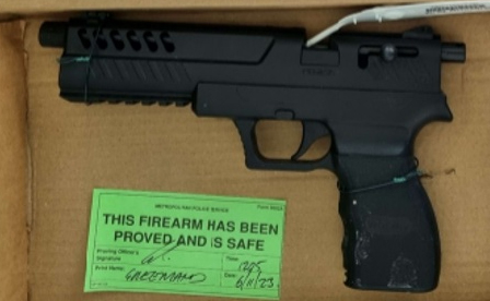 A black handgun in a box with a green tag stating "THIS FIREARM HAS BEEN PROVED AND IS SAFE".