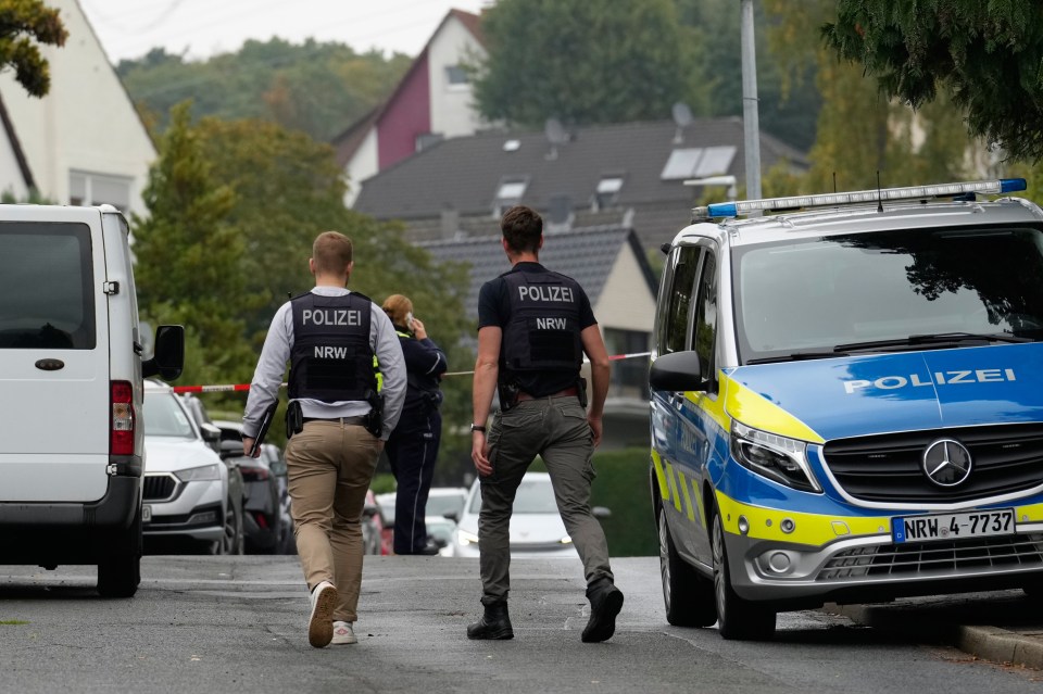 Two police officers in tactical vests with "POLIZEI" written on them, walking past a parked police van in Herdecke, Germany.