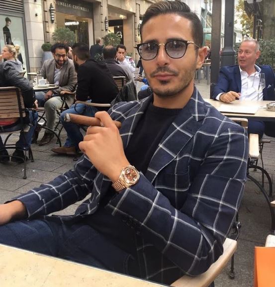 Simon Leviev (the "Tinder Swindler") sitting at an outdoor cafe holding a cigar.
