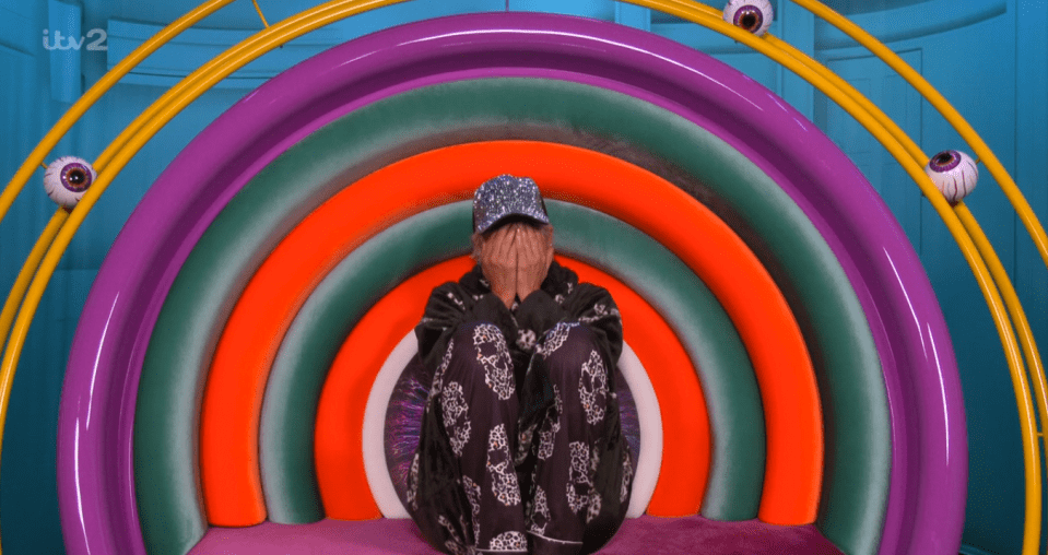 A person in a sparkly baseball cap covers their face with their hands, seated in a rainbow-colored chair.