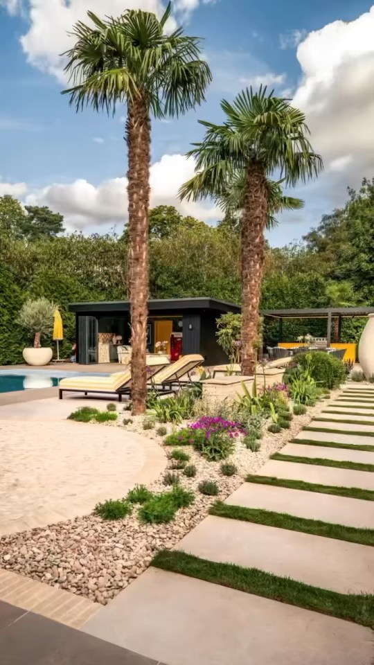 An outdoor living area with a pool, two palm trees, a dark structure with an outdoor kitchen, and a winding stone path with plantings.