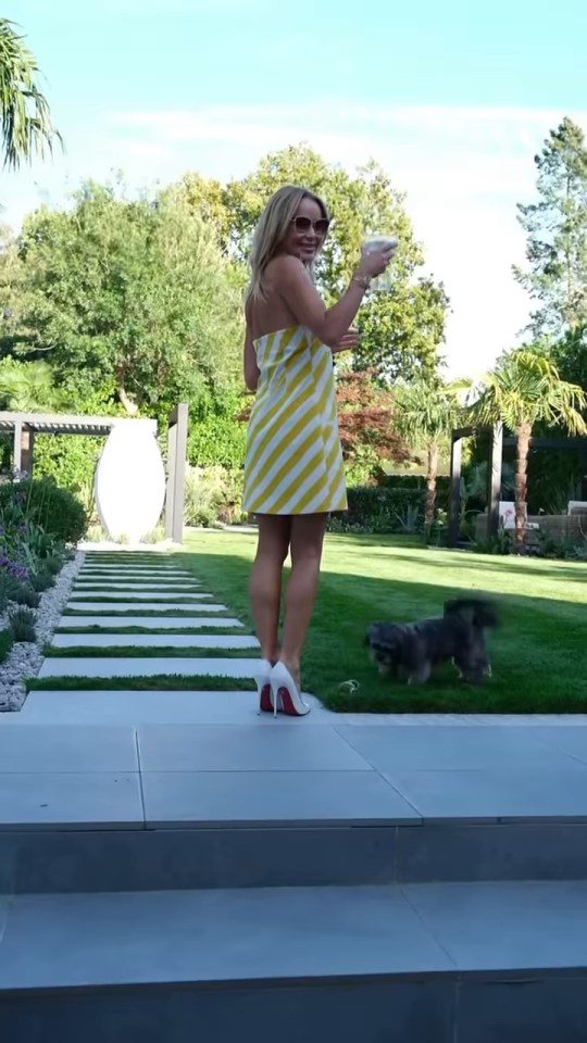 Amanda Holden in a yellow and white striped dress standing in her garden with a dog playing on the grass.