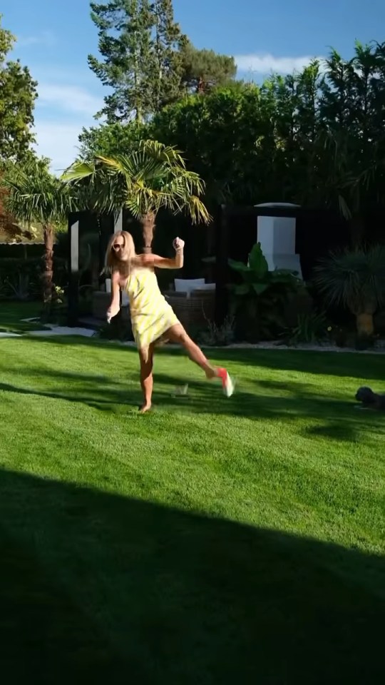 Amanda Holden barefoot in a yellow dress, kicking off a shoe while dancing on a grassy lawn.