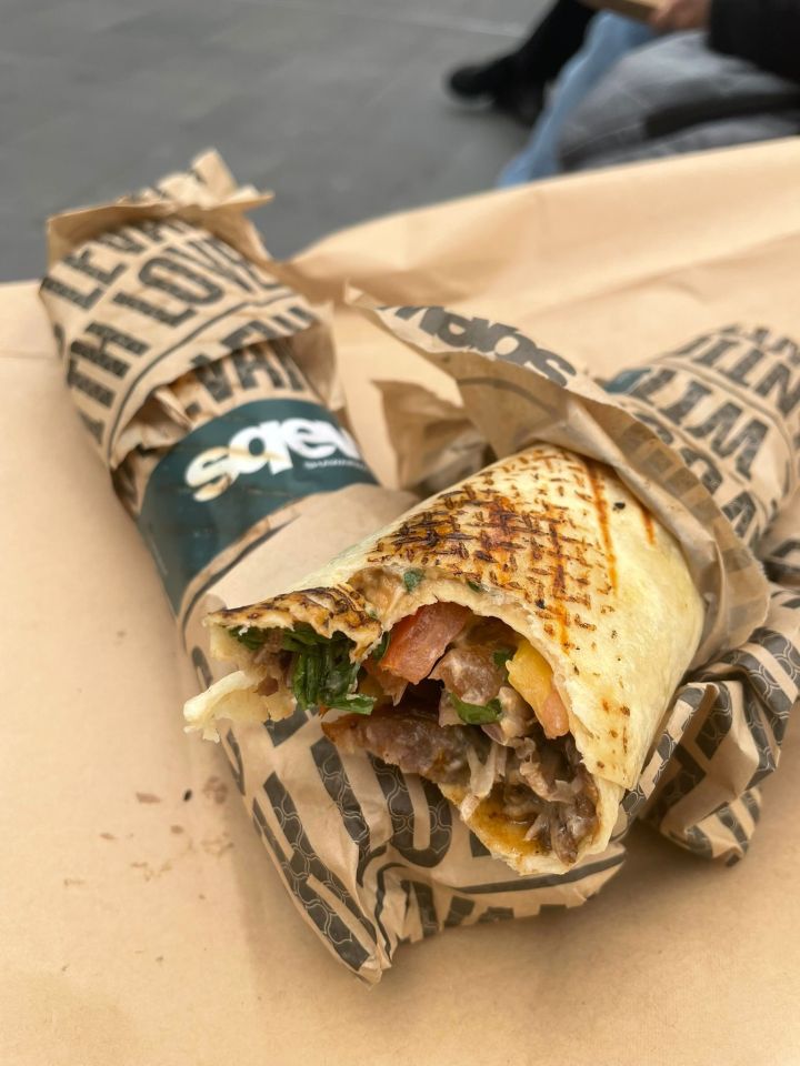 A Sqew kebab wrapped in patterned paper, with one end open showing the filling of meat, vegetables, and sauce.