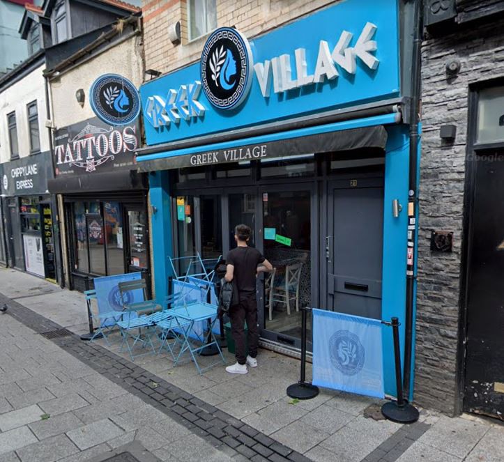 Exterior of the Greek Village restaurant with blue tables and chairs outside.