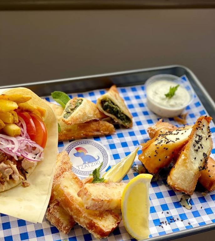 A Greek food platter from Tzatziki featuring a gyro, spinach pies, fried cheese drizzled with honey and sesame seeds, and a side of tzatziki sauce.
