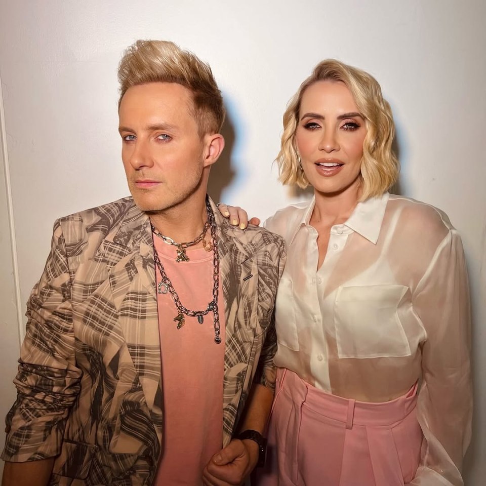 A man with blond hair and a woman with blond hair, both with pale skin, stand together against a white wall.