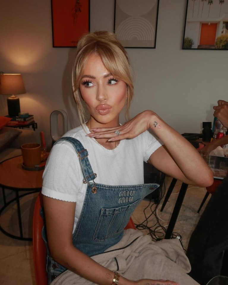 A woman with blonde hair in a bun, wearing a white shirt and denim overalls, looks to the left with pursed lips and her hand under her chin.
